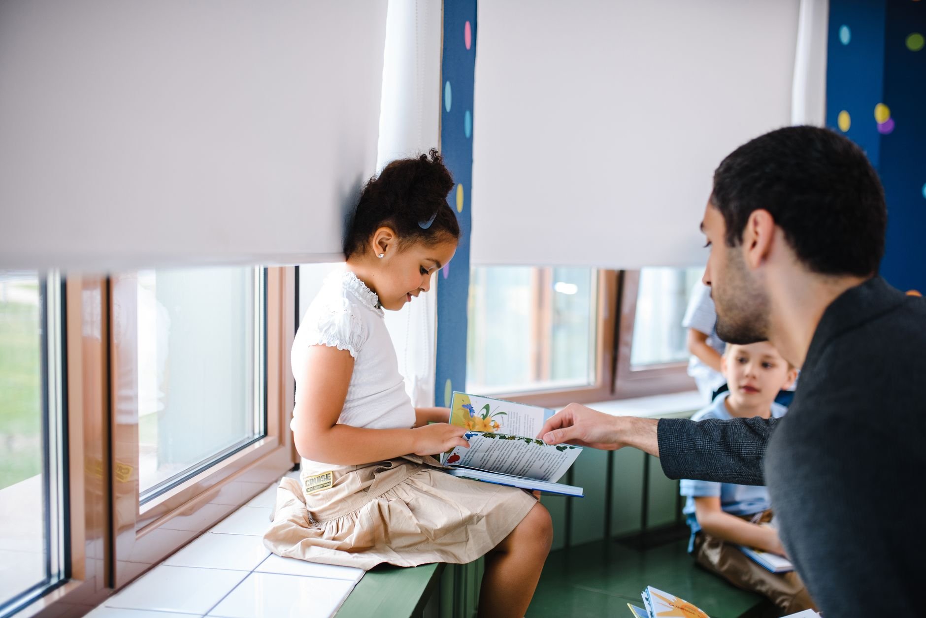 A teacher helps a young girl read a book with fellow students in a classroom setting.